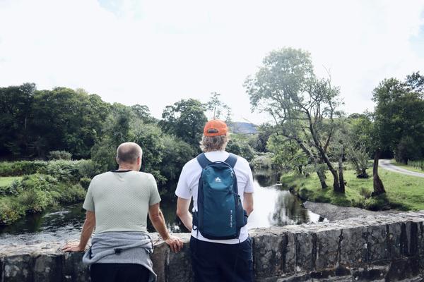 Two individuals stand on a stone bridge, their backs to the viewer, gazing out at a tranquil river flowing through a lush green landscape